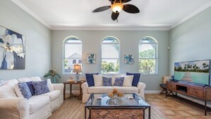A living room with a white sofa, a glass coffee table, and a TV on a wooden stand. There are three windows with blinds, a ceiling fan, and a painting on the wall.