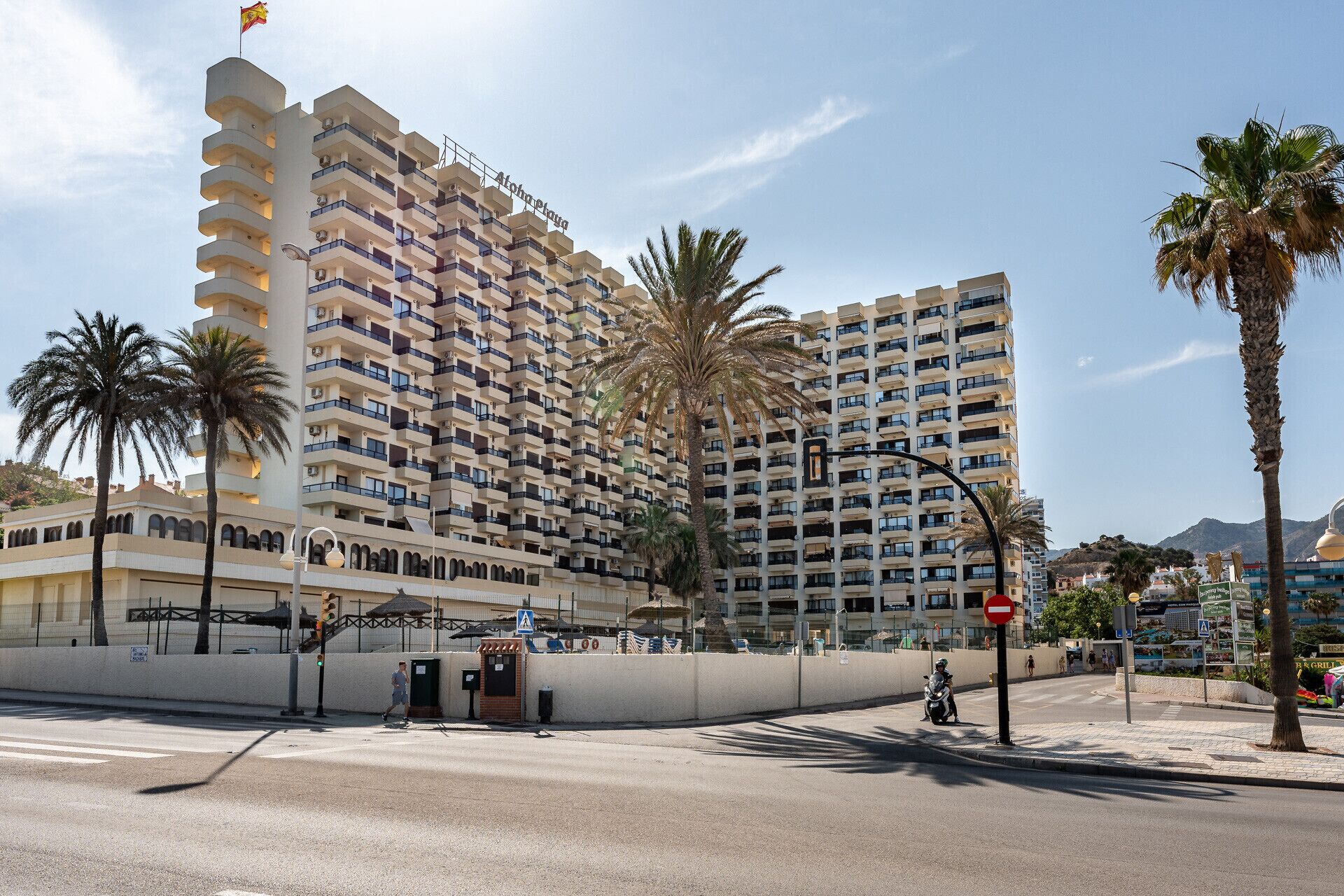 Un hotel moderno de gran altura con varios balcones, palmeras y un cielo despejado.