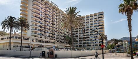 Un hotel moderno de gran altura con varios balcones, palmeras y un cielo despejado.