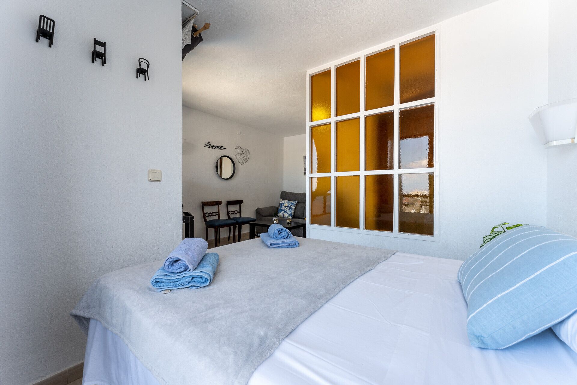 A bedroom with a large window, a bed with two rolled towels, a small table, and a chair.