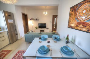 Een moderne woonkamer met een eettafel gedekt voor vier personen, een flatscreen televisie, een kroonluchter en een groot decoratief plafondschilderij.