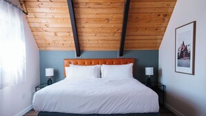 Une chambre avec un plafond en bois, un grand lit avec une literie blanche, deux tables de chevet avec des lampes et un tableau encadré au mur.