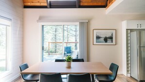 Un coin repas avec une table et des chaises en bois, une plante en pot et une vue sur une terrasse avec une chaise et un coussin bleu.