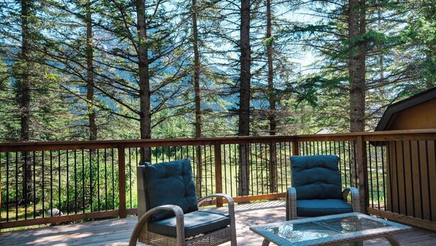 Une terrasse en bois avec deux fauteuils en osier et une table en verre, entourée de grands arbres et avec une montagne en arrière-plan.