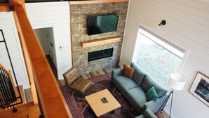 Un salon moderne doté d’une cheminée en pierre, d’un téléviseur fixé au mur et d’un coin salon confortable avec un canapé et un pouf.