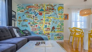 Un salon avec une fresque murale représentant une carte, un canapé gris, une table basse et des chaises jaunes.