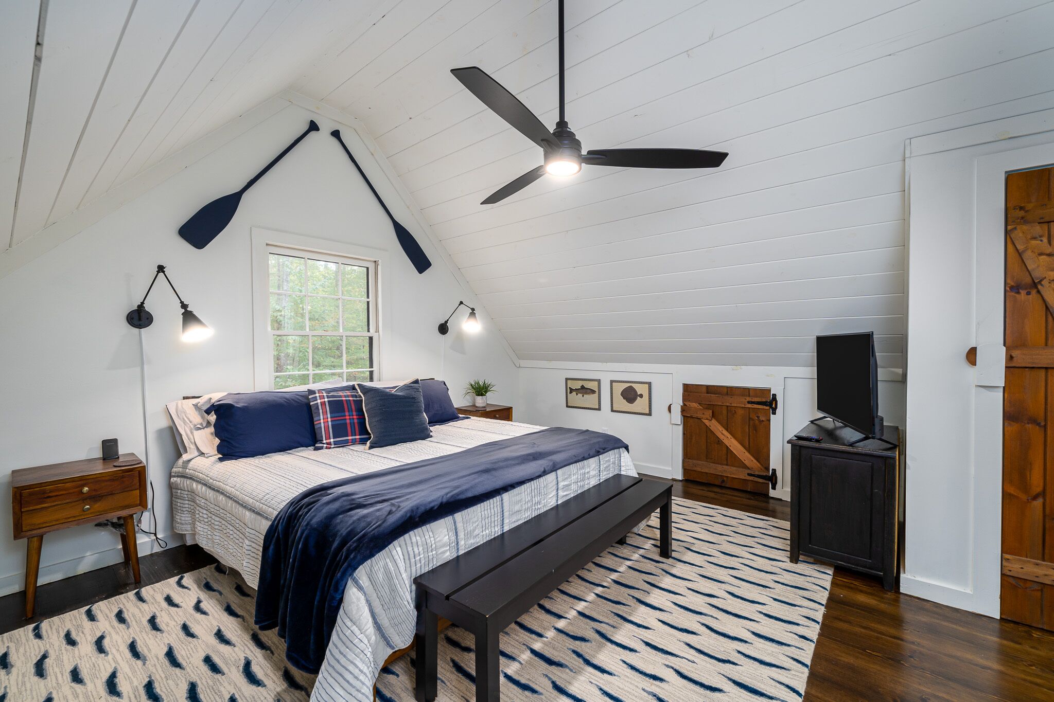 A bedroom with a sloped ceiling, a bed with a dark blue bedspread, a bench, a wooden cabinet, and a television on a stand.