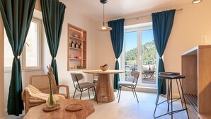 Una stanza accogliente con un tavolo da pranzo in legno, due sedie, una poltrona in vimini, una libreria e vista sull'esterno.