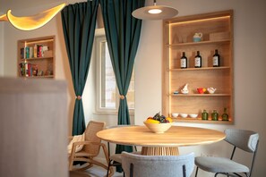 A dining area with a round wooden table, two chairs, a bowl of fruit, a bookshelf, and a shelf with bottles.
