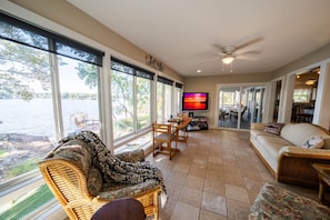 A spacious living room with large windows, a television, wicker furniture, and a view of a lake.