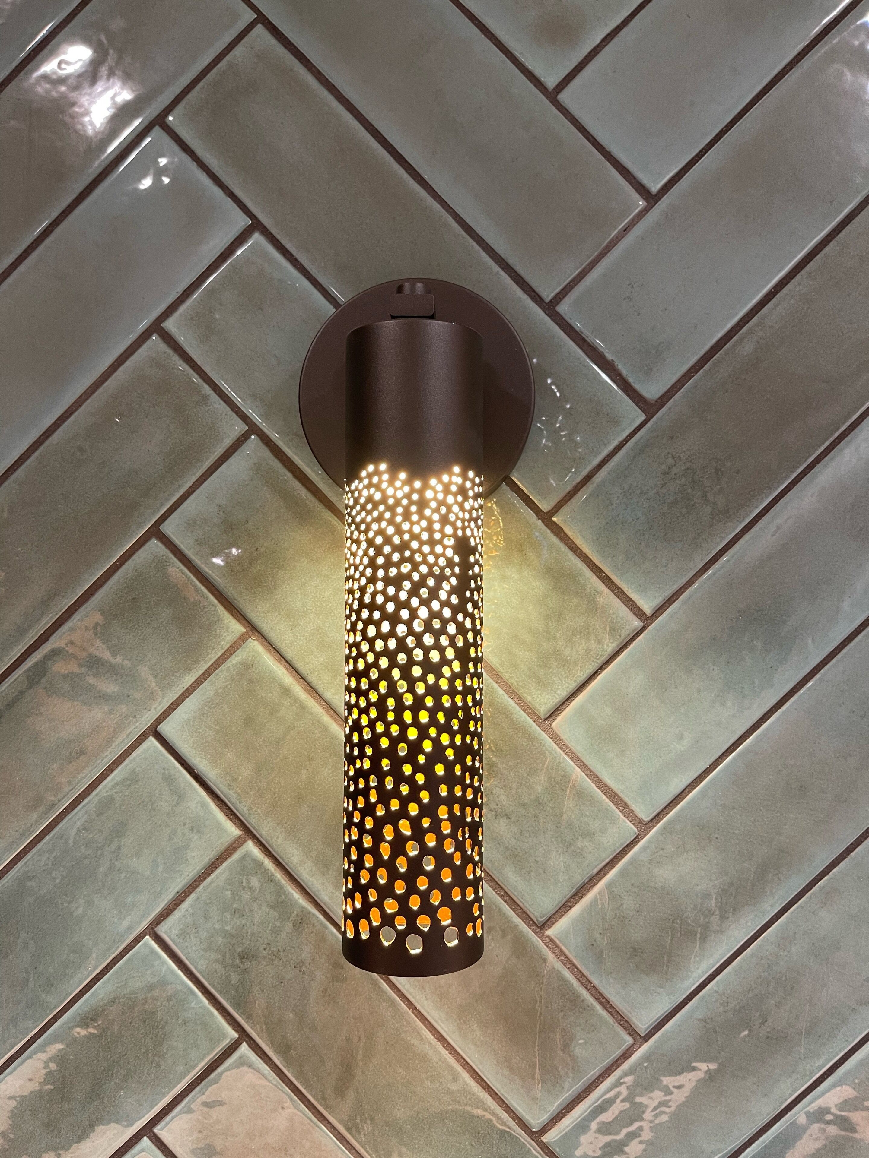 Una lámpara de pared con forma cilíndrica y diseño perforado, sobre un suelo de baldosas con patrón de espiga.