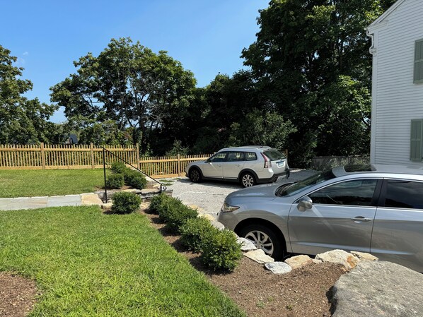 Un camino de entrada residencial con dos coches aparcados, una valla de madera y una casa de fachada blanca.