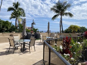 An outdoor patio with tables and chairs, surrounded by palm trees and potted plants. - Condo in downtown Kailua-Kona - walk to restauants, shops, the beach (Kailua Kona)