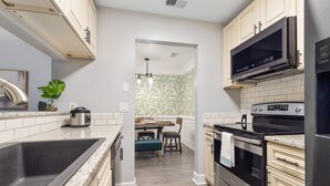 Cocina moderna con pileta de acero inoxidable, microondas y horno. Hay un comedor con mesa y sillas, y una planta en la encimera.