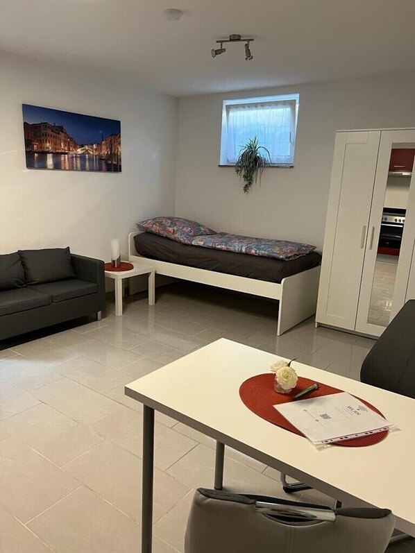 A compact living space with a sofa, a small table with a red placemat, a bed with a colorful mattress, a window with a plant, and a kitchenette in the background.