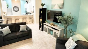 Een moderne woonkamer met een donkergroene bank, een salontafel en een televisie op een dressoir. Achtergrond toont een keuken met een bar en barkrukken.