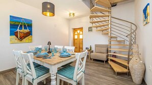 Un comedor con una mesa de madera dispuesta para cuatro personas, rodeada de sillas blancas con cojines color turquesa. Se observa una escalera de caracol con escalones de madera y barandillas de metal a la derecha.