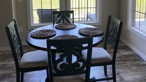 A dining area with a round table, four chairs, and two windows with sheer curtains.