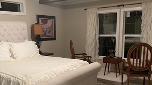 A bedroom with a large bed, wooden floor, a chair, a table, and a window with curtains.