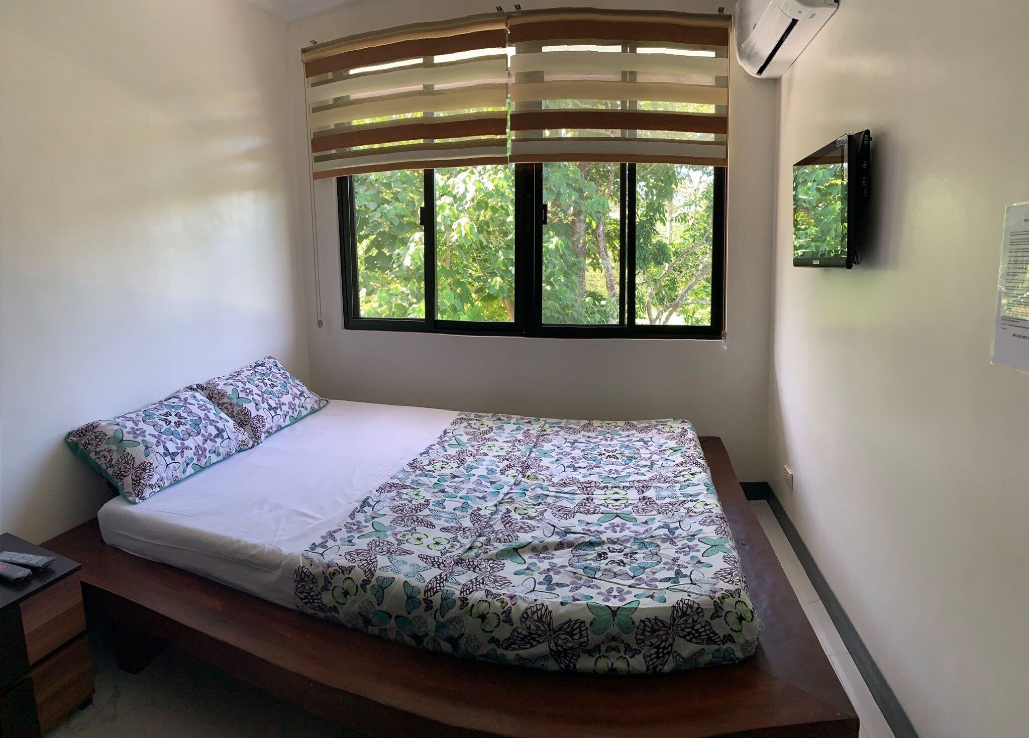 A single bed with a floral bedspread, a bedside table with a remote control, and a window with blinds.