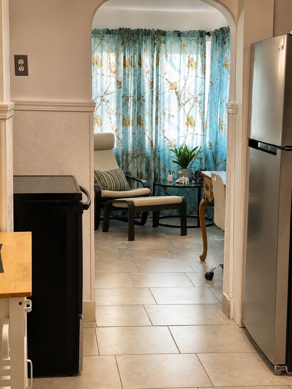 A kitchen with a refrigerator, a trash bin, a dining table with chairs, and a window with curtains.