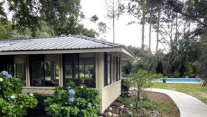 Ein Haus mit überdachter Veranda, einem Swimmingpool und einem Garten mit verschiedenen Pflanzen und einer kleinen Statue.