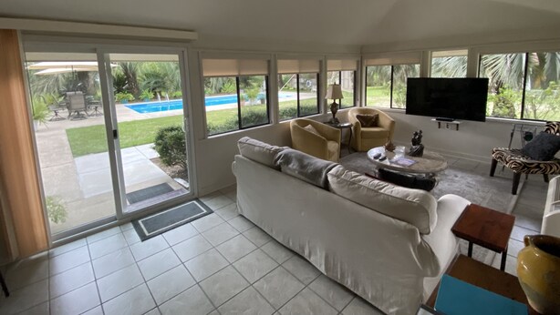 Ein großzügiger Wohnraum mit einer großen Couch, einem Couchtisch und einem an der Wand montierten Fernseher. Eine Glastür führt auf eine Terrasse mit Pool und Gartenmöbeln.