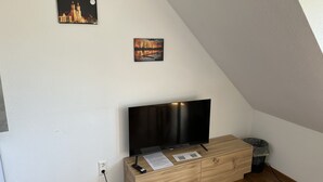 Huoneessa on televisio puisen kaapin päällä, roskakori ja kaksi kehystettyä kuvaa seinällä.