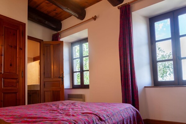 Un dormitorio con techo de madera, cama con colcha estampada, ventana con vista a árboles y puerta que conduce a otra habitación.