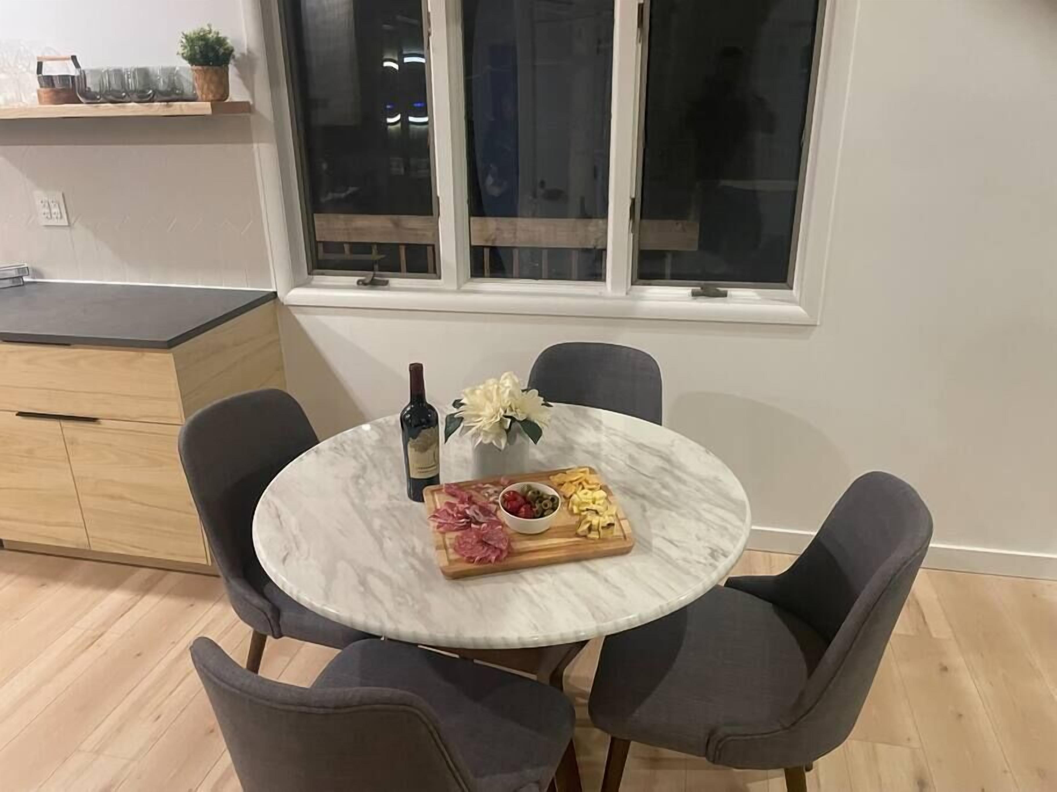 A round marble table with four chairs, a bottle of wine, a floral centerpiece, and a charcuterie board.