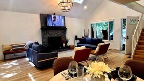 Una sala de estar moderna con chimenea de piedra, televisor montado encima y comedor con copas de vino y un florero.