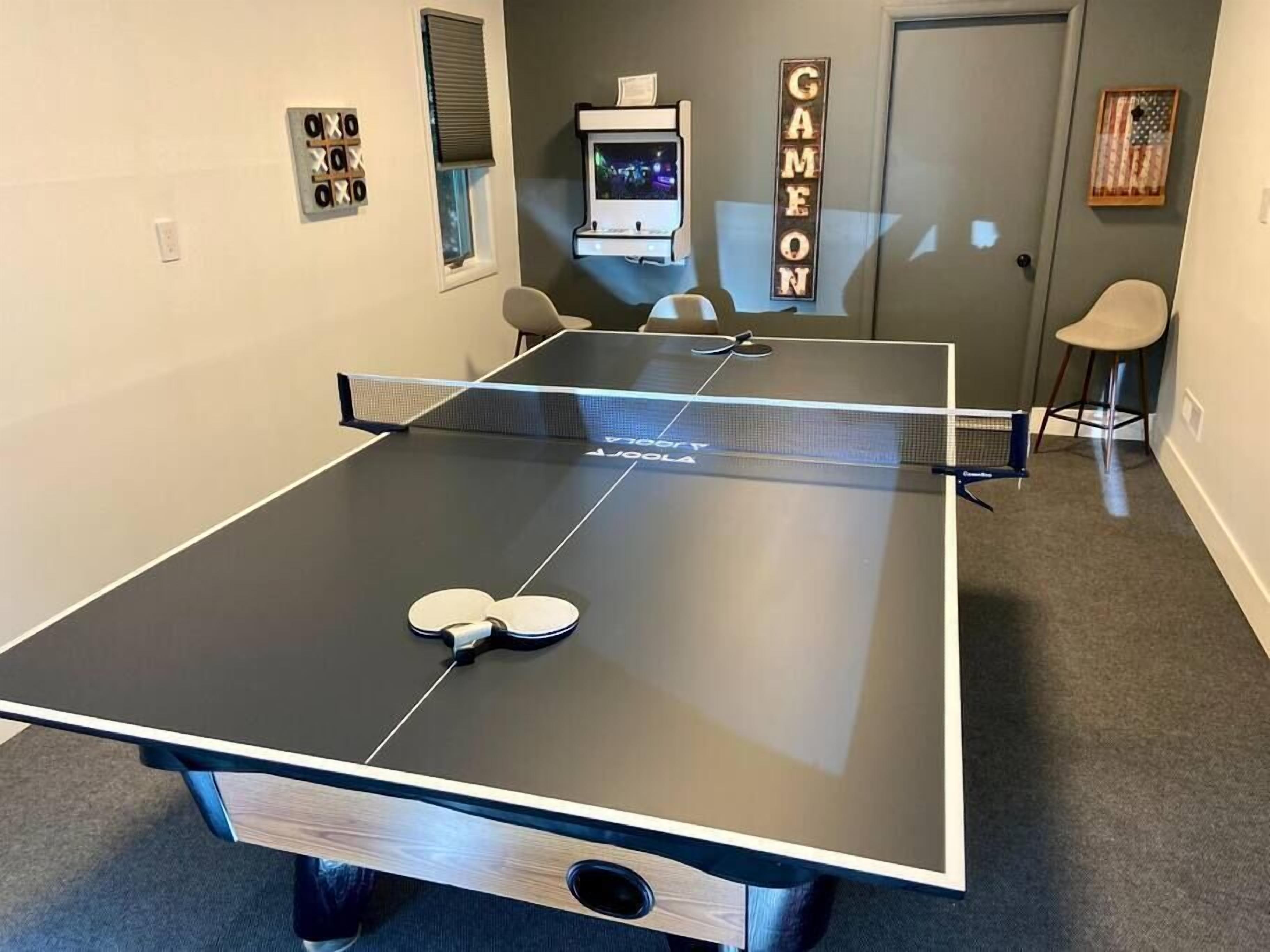 A ping pong table with two paddles, a TV, and a 'GAME ON' sign in a room with a door, window, and chairs.