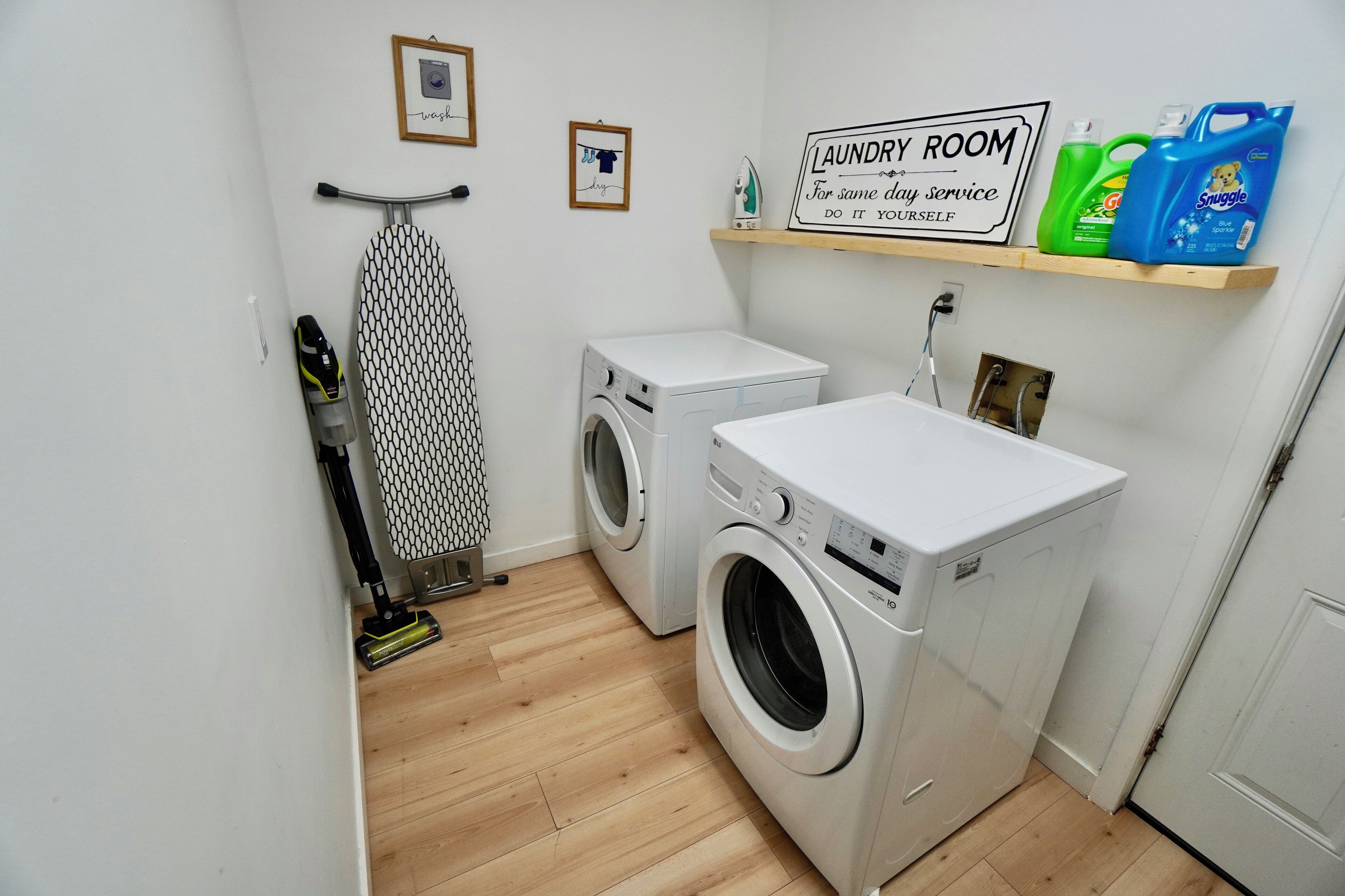A laundry room with two washing machines, an ironing board, and a shelf with laundry supplies.