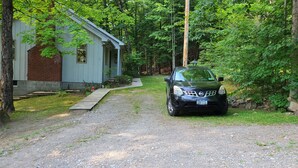 A gravel driveway leading to a small, light blue building with a brick chimney, surrounded by dense green foliage. -  Dog friendly lakefront home w/fireplace near Lk George. Telecommuters welcome! (Fort Ann)