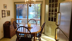 A dining area with a wooden table and chairs, a chandelier, and a view of a balcony with trees and a pool. -  Dog friendly lakefront home w/fireplace near Lk George. Telecommuters welcome! (Fort Ann)