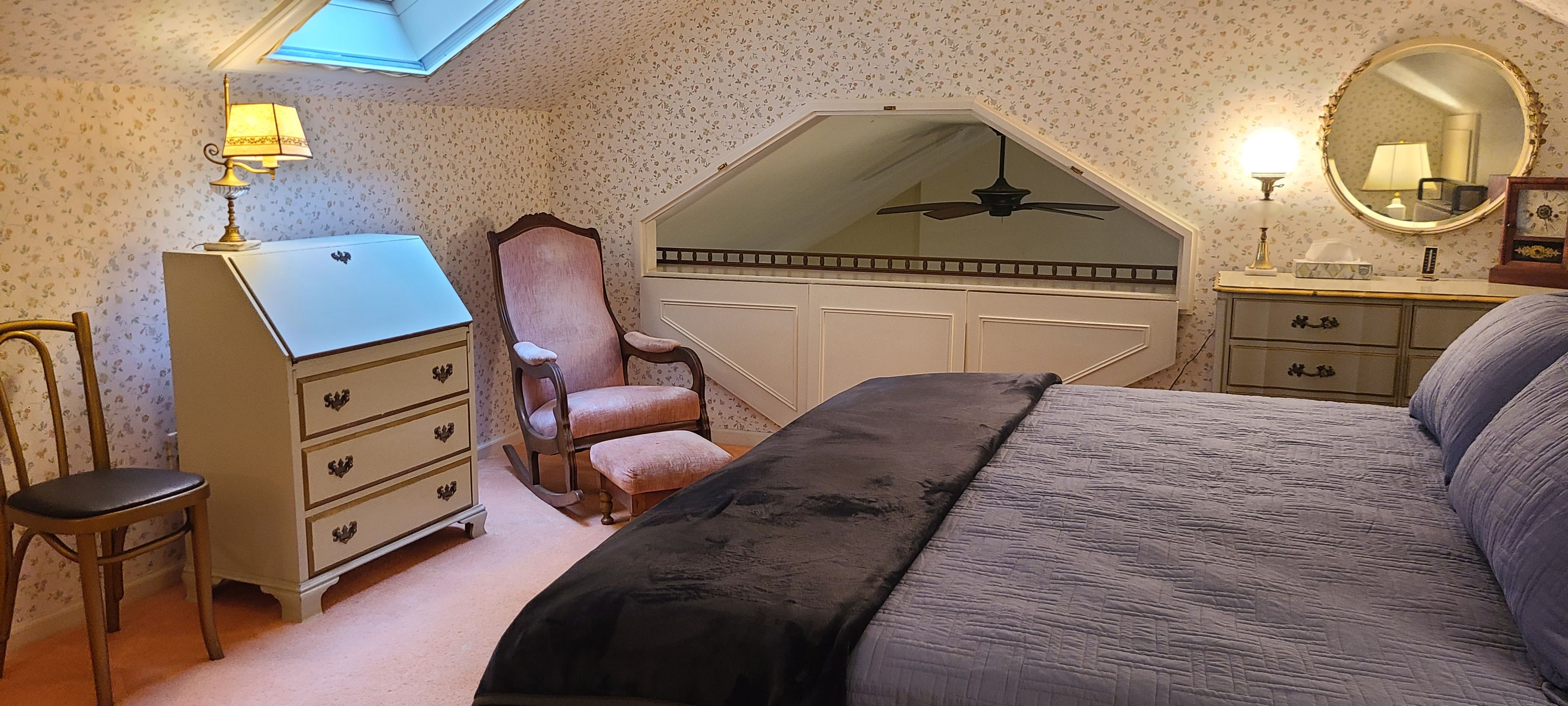 Un dormitorio con techo inclinado, cama, cómoda, silla y espejo.