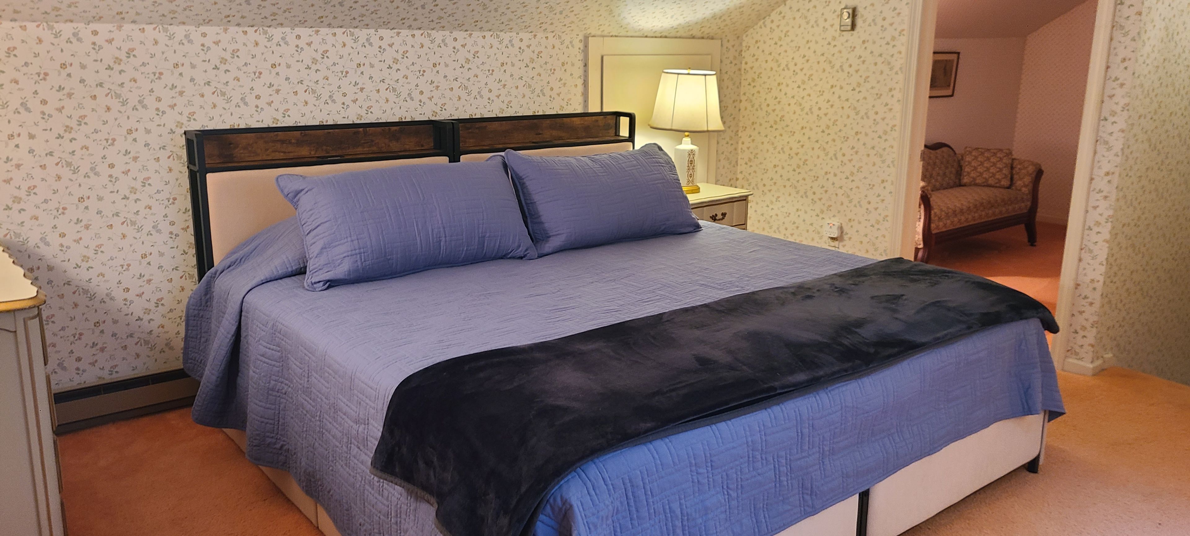Una cama bien hecha con cabecera, una mesita de noche con lámpara y una silla al fondo.