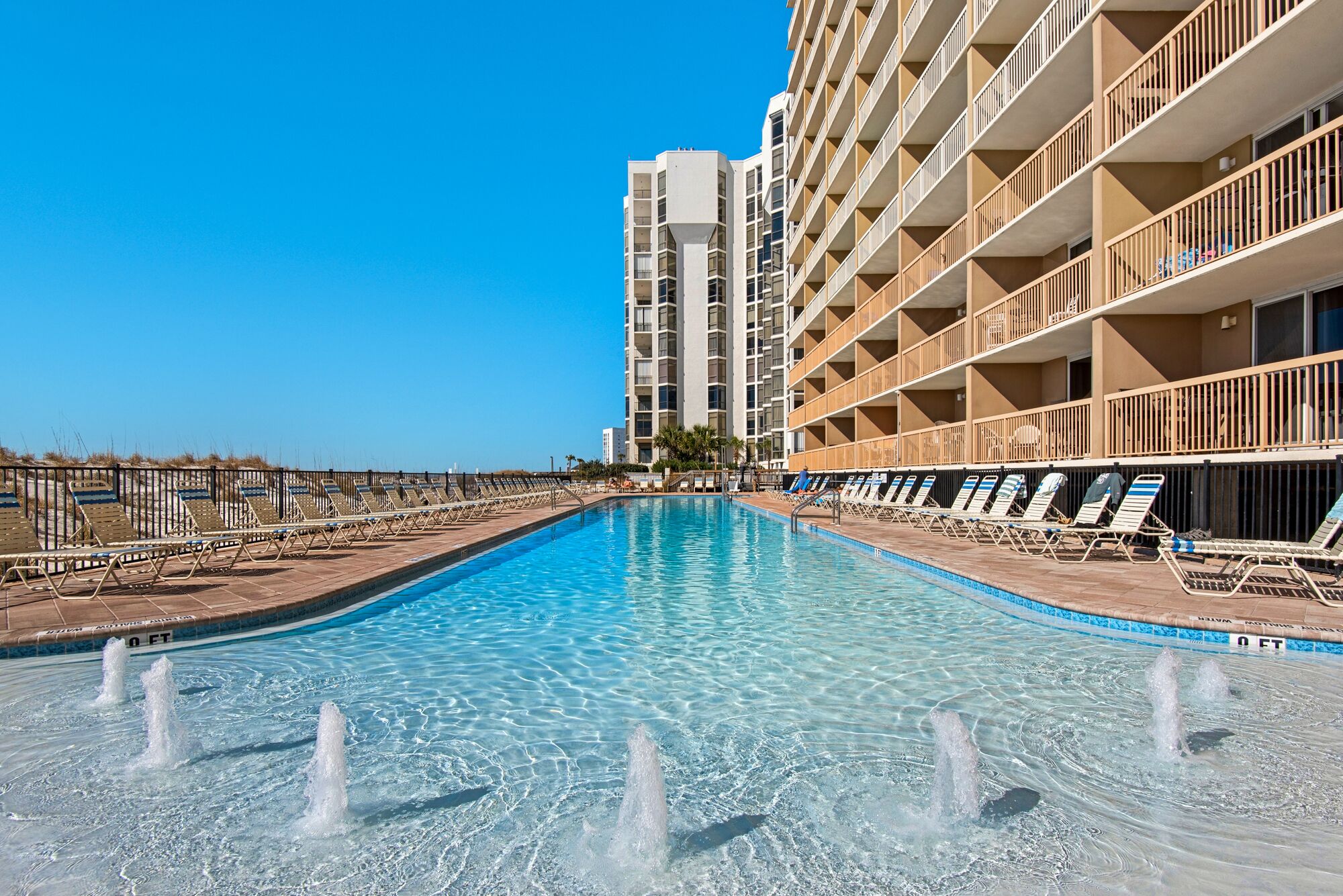 A swimming pool with water fountains, surrounded by lounge chairs and a high-rise building with balconies.