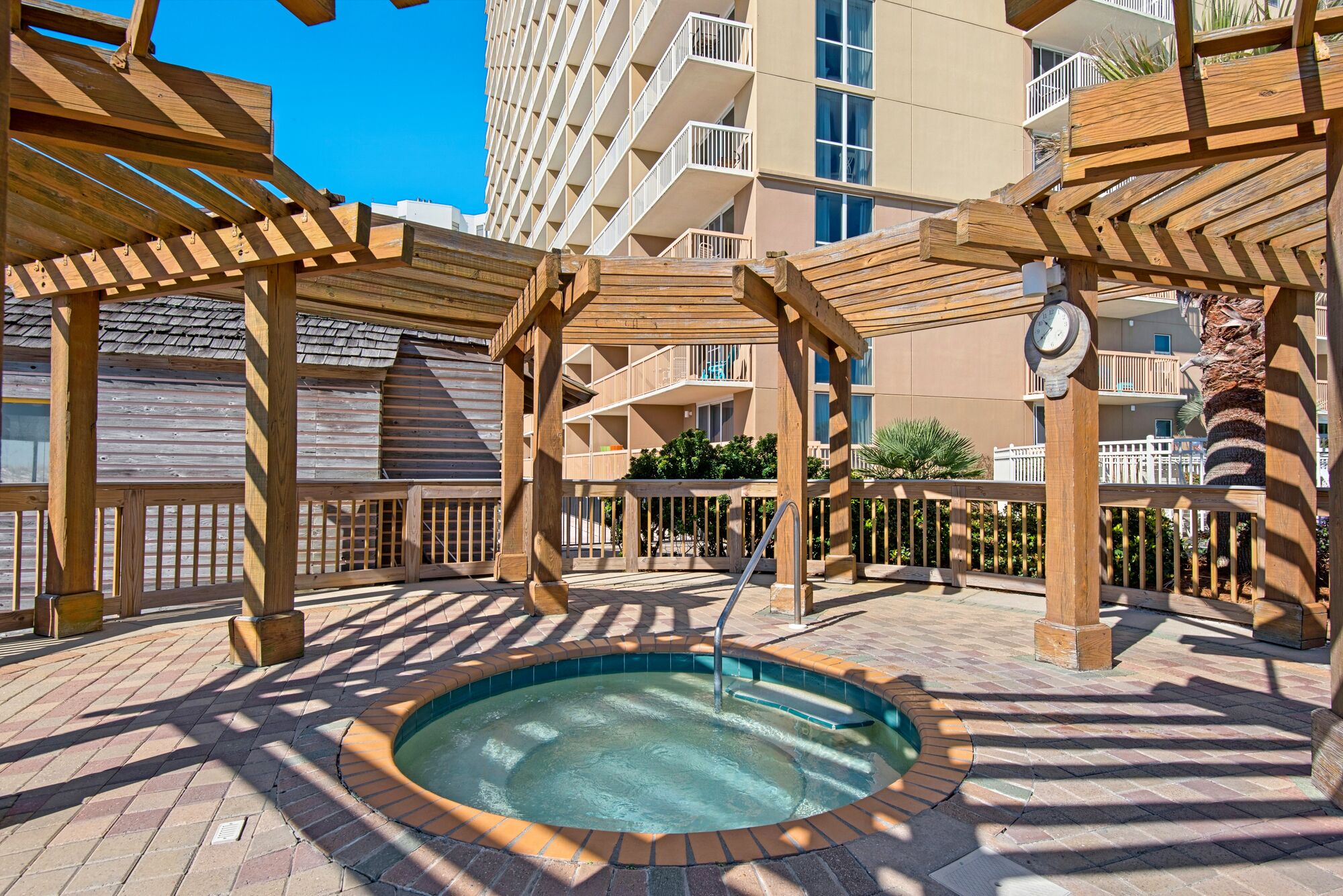 Um deck de madeira com uma banheira de hidromassagem, cercado por uma piscina, e um edifício com varandas ao fundo.