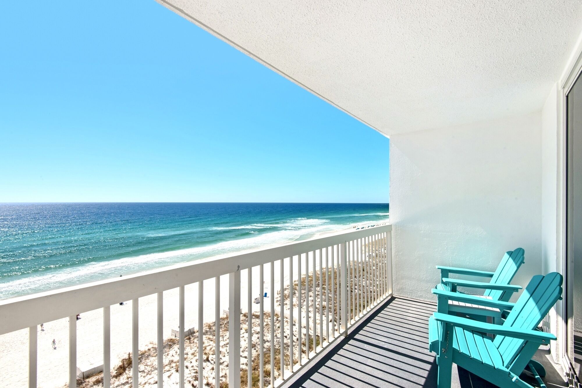 A balcony with a view of the ocean, a white railing, and two turquoise chairs.