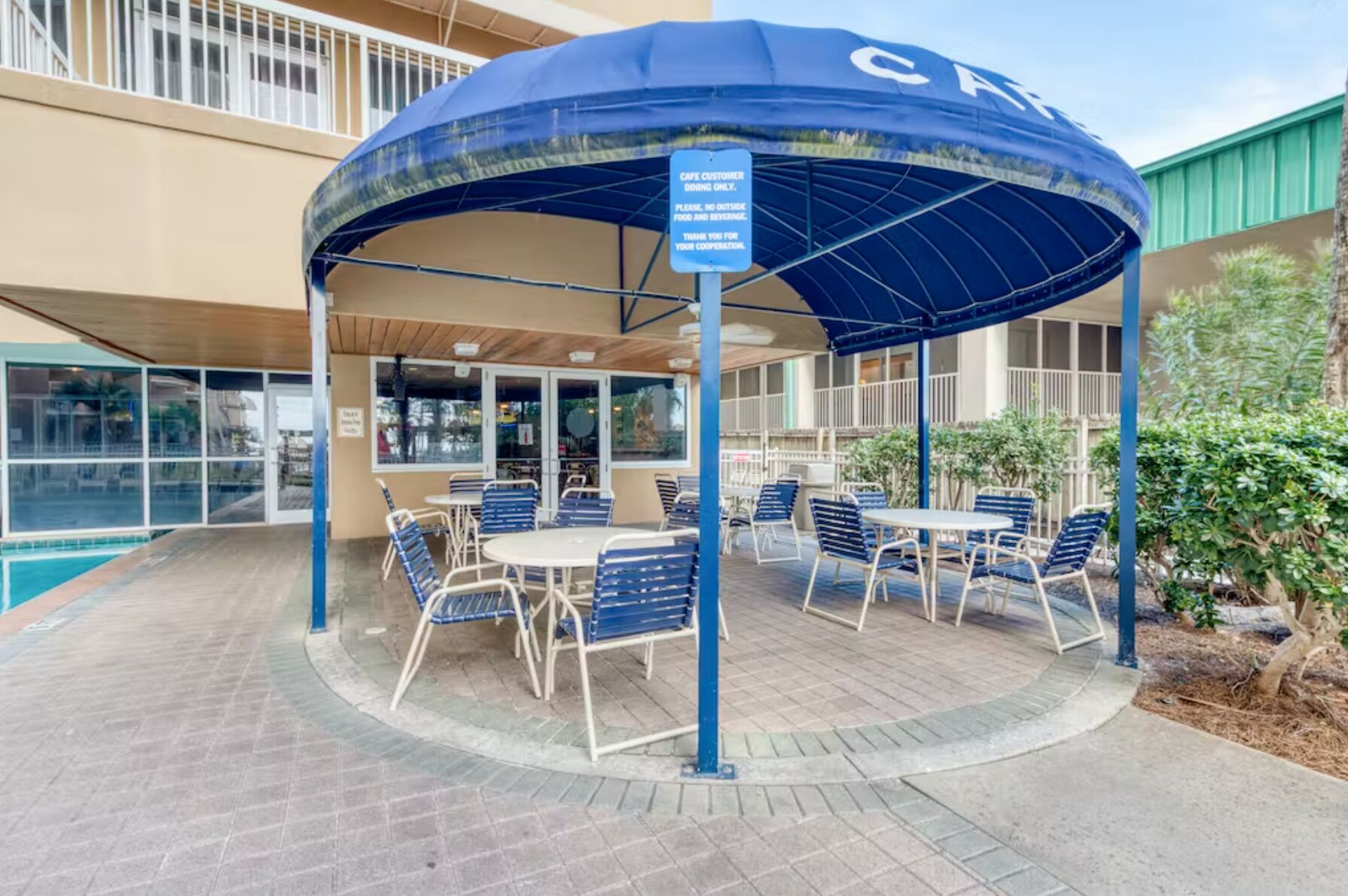 A covered outdoor seating area with tables and chairs, a sign indicating it is for cafe customers only, and a pool in the background.