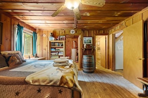 A cozy cabin interior with a bed, a bookshelf, a wooden barrel, and a door leading to another room.