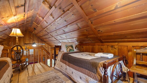 A wooden cabin bedroom with a bed, a lamp, a rocking chair, and a spinning wheel. - Quaint, cozy vacation cabin with hot tub near Natural Bridge State Park (Rogers)