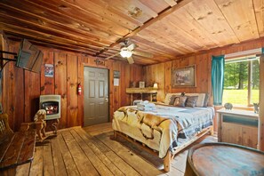 A cabin interior with a bed, a fireplace, a table, and a window with a view of the outdoors. - Quaint, cozy vacation cabin with hot tub near Natural Bridge State Park (Rogers)