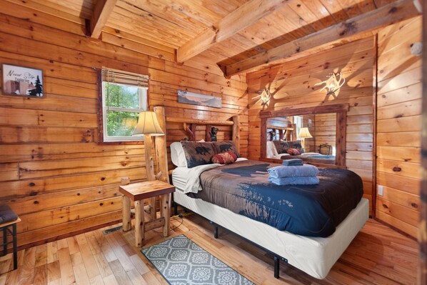 Un dormitorio rústico de cabaña de troncos con una cama, una mesita de noche, una lámpara y una ventana con vistas a la vegetación.