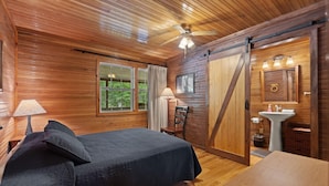 Een houten slaapkamer met een bed, een bureau, een stoel, een plafondventilator, een raam met gordijnen en een schuifdeur naar een badkamer.