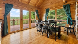 A wooden cabin with a dining area featuring a table and chairs, large windows with curtains, and a view of a wooden deck and greenery. - Large Luxury Log Cabin, Sleeps-13, Spectacular view! Loaded with ammenities! (Rogers)