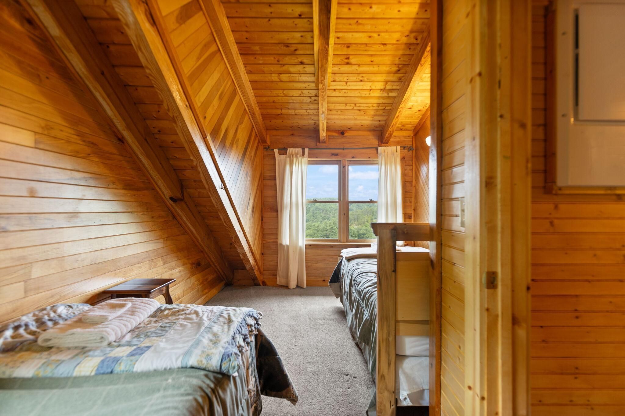 A cozy attic bedroom with wooden walls and a sloped ceiling, featuring two beds and a window with curtains.