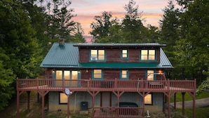 A two-story wooden cabin with a green roof, a covered porch, and a small deck. - Large Luxury Log Cabin, Sleeps-13, Spectacular view! Loaded with ammenities! (Rogers)