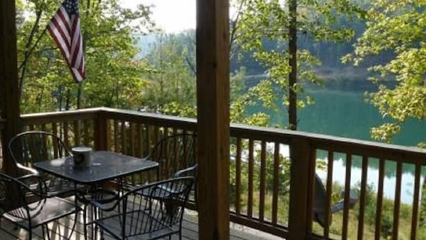 Een houten terras met een tafel en stoelen, een Amerikaanse vlag en uitzicht op een meer, omgeven door bomen.